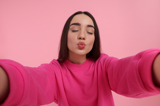 Young woman taking selfie and sending air kiss on pink background