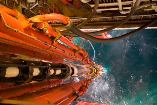 offshore oil rig riser pipe view to sea through moonpool viewed from height. Main pipe contains drilling fluids and drill pipe and connects to the seabed wellhead equipment. Mud line hoses and hydraulic tensioners also seen.