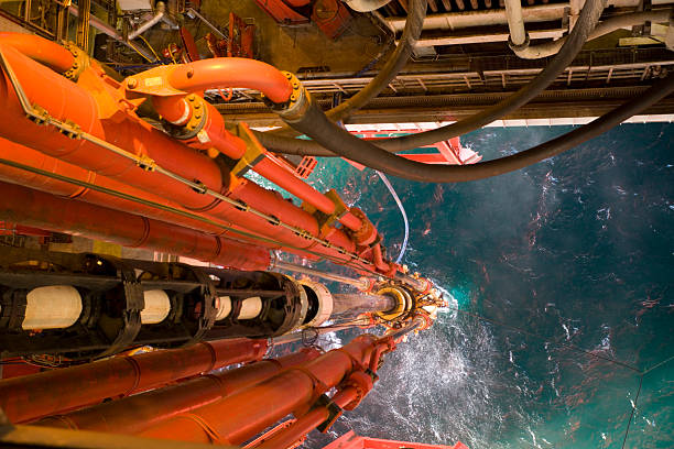 oil rig view riser pipes down to sea level offshore oil rig riser pipe view to sea through moonpool viewed from height. Main pipe contains drilling fluids and drill pipe and connects to the seabed wellhead equipment. Mud line hoses and hydraulic tensioners also seen. oil industry stock pictures, royalty-free photos & images