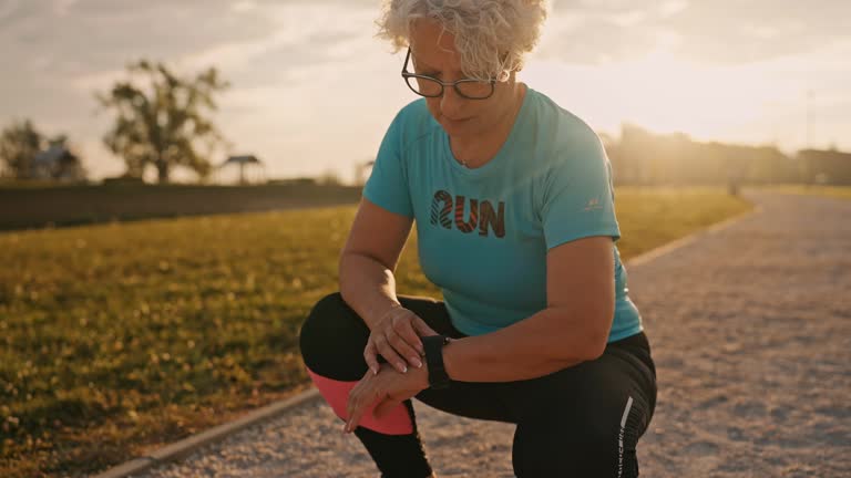 SLO MO Senior Woman in Sportswear Looking at Smartwatch to Check Heart Rate after Finishing Jogging in Park at Sunset