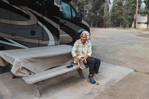 An active and healthy senior woman of Chinese and Hawaiian descent sits at a picnic table outside her RV which is parked at a campground in Oregon and puts on her shoes.