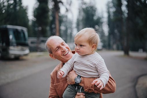 An active and healthy senior man affectionately carries and looks at his adorable one year old Eurasian grandson while on a leisurely walk through a campground in Oregon.
