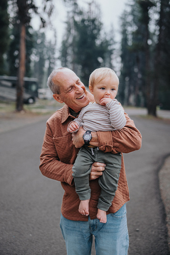 An active and healthy senior man affectionately carries and looks at his adorable one year old Eurasian grandson while on a leisurely walk through a campground in Oregon.