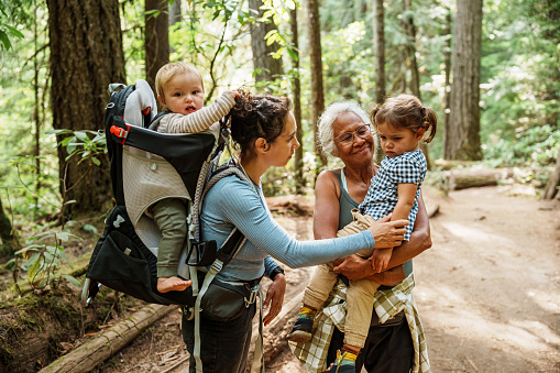 A vibrant senior woman of Pacific Islander descent who is hiking in a lush forest with her multi-generation family affectionately holds her three year old granddaughter as the child's mom comforts the sad little girl who fell down and got hurt while hiking.
