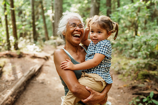 A healthy and adventurous senior woman of Hawaiian and Chinese descent laughs joyfully while carrying her adorable three year old Eurasian granddaughter during a hike through a lush forest in Oregon.