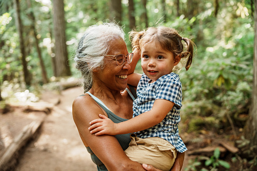 A healthy and adventurous senior woman of Hawaiian and Chinese descent laughs joyfully while carrying her adorable three year old Eurasian granddaughter during a hike through a lush forest in Oregon.