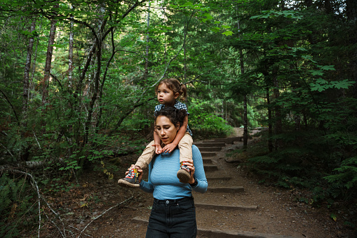 An active Eurasian mom hikes through a lush mountain forest in Oregon while carrying her three year old daughter on her shoulders.