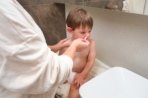 The mom is washing her baby nose with a cotton swab in the bathroom, as part of their daily hygiene routine. Kid aged two years (two-year-old boy)