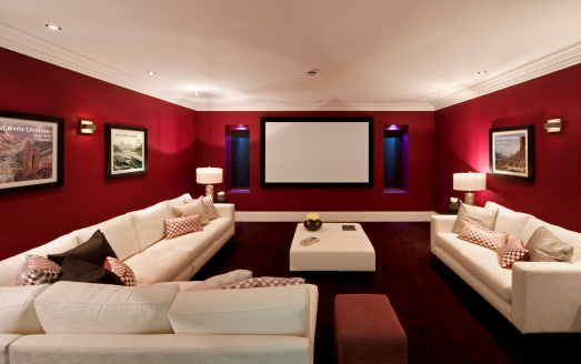 a cinema room in an expensive new home with large comfortable cream coloured settees, silk cushions and a foot stool. On either side of the screen are recessed windows with electronic blinds (lowered) and alcove lights. The pictures on the wall are the photographer's.