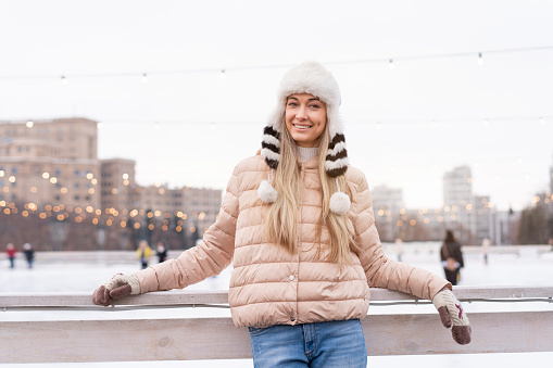 Beautiful lovely middle-aged girl blonde hair warm winter jackets knitted glove stands ice rink background Town Square. Christmas mood lifestyle Happy holiday woman walk snowy day Winter leisure