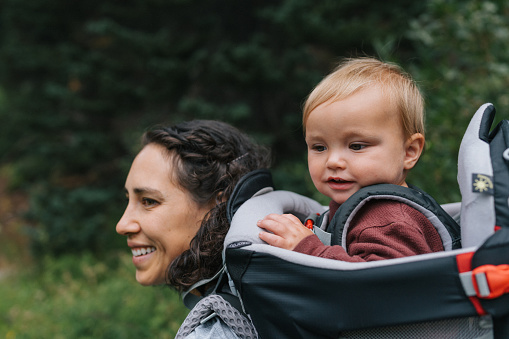 Side profile of a young mom of Eurasian ethnicity smiling, carrying her one year old son on her back as they hike through a trail near Crater Lake in the Oregon summertime.