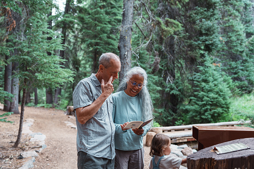 A multiracial senior couple stands with their Eurasian three year old granddaughter, studying a map while taking a break from a family hike in the Oregon summertime.