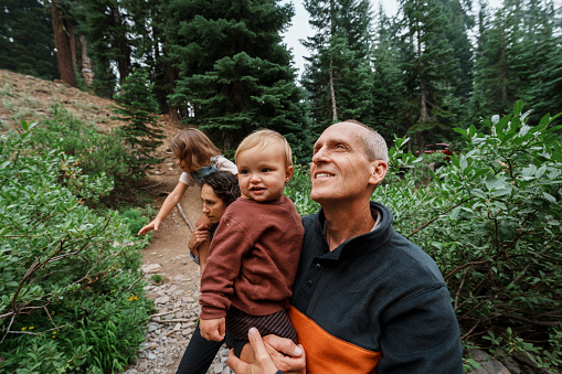 Caucasian senior man holds his multiracial one year old grandson, looking around at the nature surrounding them while hiking together in Oregon.