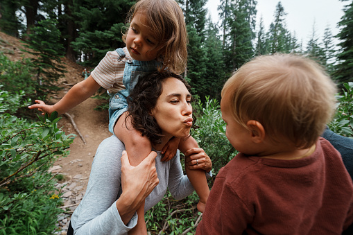 Loving mother of Eurasian ethnicities leans in to kiss her one year old son, while holding her preschool age daughter on her shoulders during a family hike in the Oregon forest.