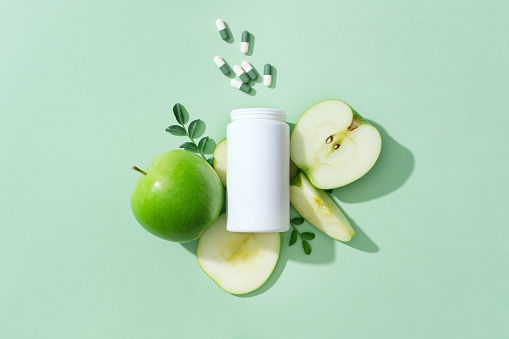 On the pastel background, a white medicine bottle unlabeled with capsule pills decorated with fresh slices of green apple. Advertising photo for product with natural extract for weight loss