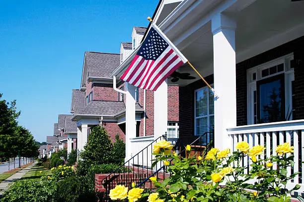 Detail of a cottage-style front porch with an American Flag hanging from the front column. A row of homes fade off into the background while spectacular yellow roses are in full bloom in the foreground.
