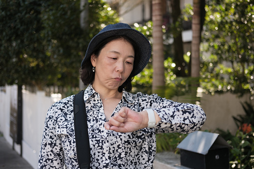 Asian woman walking along a suburban street wearing a hat and checking the time on her watch. Upset expression.