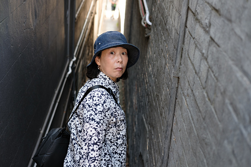 Scared middle aged Asian woman in hat and floral shirt looking over her shoulder in a narrow path between two buildings
