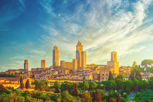 At sunrise in San Gimignano, Tuscany, Italy, the sky paints a mesmerizing canvas of yellow, green, and blue hues. As the first light of day graces the scene from the right, one side of the iconic towers and buildings glistens with a radiant golden-orange glow. This enchanting display creates a mesmerizing  and captivating atmosphere, casting a warm embrace over the historic town.