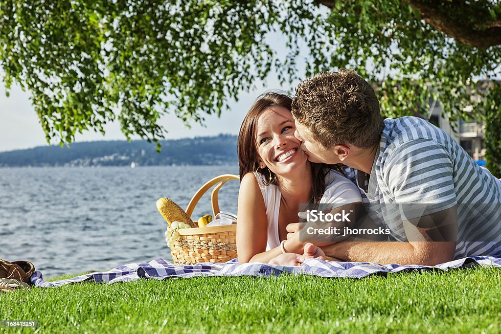 Young Couple on Romantic Picnic Photo of an attractive young couple enjoying a warm summer day with a picnic at the park, boyfriend kissing girlfriend on the cheek. Lake Stock Photo