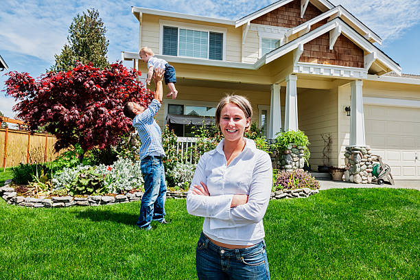 Proud Mother at Home with Family Photo of a happy family in front of their home, mother in foreground while father plays with son in the background. in front of stock pictures, royalty-free photos & images