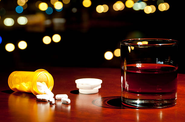 Whisky or Rum and a bottle of spilled pills Glass of Whisky or Rum next to a spilled bottle of pills on a wooden table, with city lights in the background. cirrhosis stock pictures, royalty-free photos & images