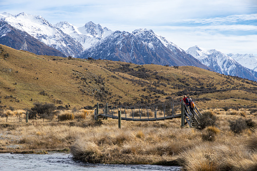 grassland area with snowy mountain range in the background with two people crossing a bridge over a river