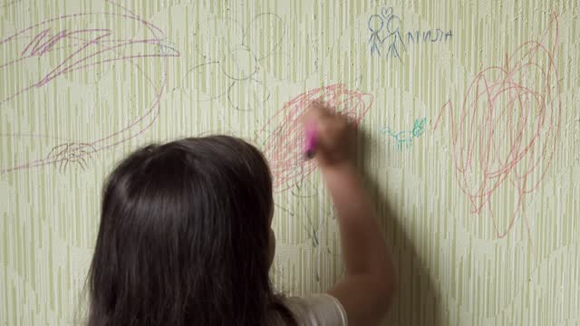 Black haired little girl draw doodle with multi colored marker on wallpaper back view, develop artistic skills. Space for painting, disorderly conduct, play pranks, paperhanging damage