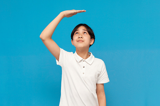 asian boy of twelve years old measures his height with his hand on blue isolated background, korean not tall child dreams of growing up and holds his hand above his head
