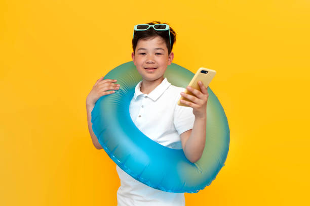 黄色の背景に膨脹可能な水泳リングを持つ10歳のアジアの小さな男の子は、スマートフォンを使用します - 10 11 years little boys glasses portrait ストックフォトと画像