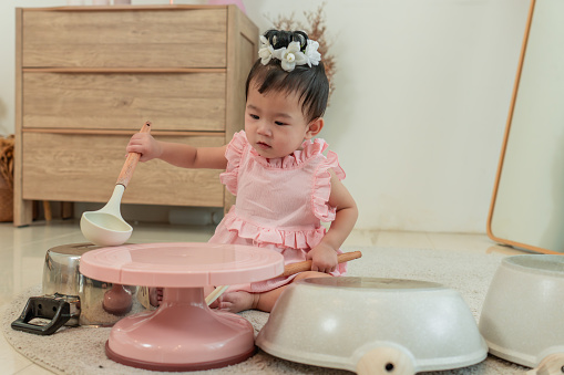 Adorable Asian little baby girl with pink dress, playing with pastel cooking station, oven, pan for cooking meal at home. Happy healthy girl having fun playing with her toy kitchen and pretending to cook breakfast.