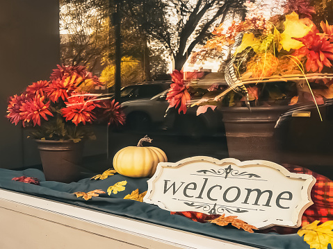 The window display of a small local business has a welcome sign in it.   The window is decorated for the autumn season. It is a generic location.  There are reflections of street parking in the window