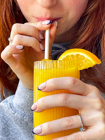 Stock photo showing close-up view of orange juice with orange slice in glass being drunk by red haired woman through paper straw at an al fresco restaurant table.