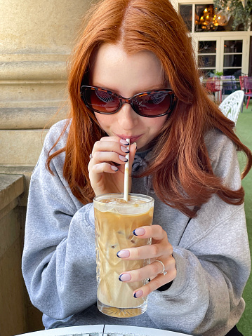 Stock photo showing close-up view of iced coffee in glass being drunk by red haired woman through paper straw at an al fresco restaurant table.
