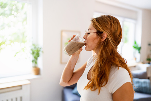 In her modern home, young redhead Caucasian woman with vitiligo, drinking healthy smoothie she made