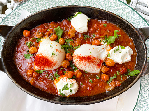 Stock photo showing close-up, elevated view of a cooking pot full of Mediterranean healthy breakfast of shakshouka / shakshuka / chakchouka. The recipe consists of chickpeas, egg poached, peppers, onion, olive oil and garlic in savoury tomato sauce spiced with cumin and paprika cayenne pepper.