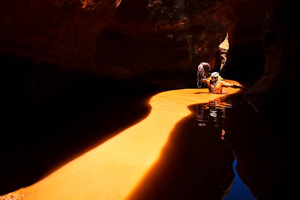 canyon adventure! a man wades through water that is illuminated by the sunlight creating an orange streak through the dark black narrows of a slot canyon.  the man holds a rope above his head used for climbing and rappelling in the slot canyon.  such beautiful nature scenery landscapes and outdoor adventure can be found in neon canyon in the grand staircase escalante national monument of utah.  horizontal composition with copy space in darkness. escalante stock pictures, royalty-free photos & images