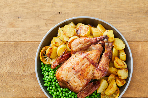 Roasted chicken with potatoes and green peas on an iron plate, with Rosemary, green peas, sea salt and pepper, served on a wooden rustic home kitchen or restaurant table, representing food culture, city life, indulgence in meat culture, healthy family protein meal, healthy eating and healthy lifestyle, wellbeing and body care