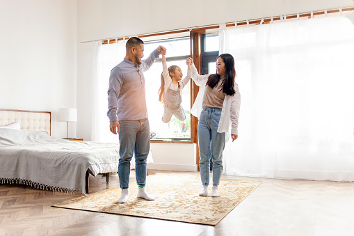 happy Asian family with little daughter stands at home in the bedroom and rejoices, dad and mom raise the hands of cheerful child in their house