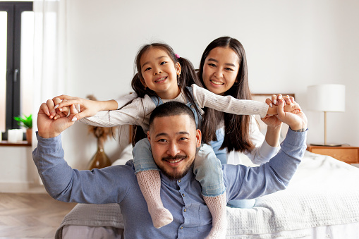 happy asian family sit together at home in the bedroom and smile, little korean girl sits on the shoulders of her dad and rejoices with her parents
