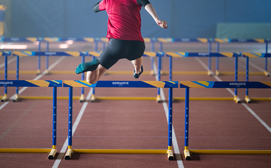 Young male runner training and jumping through hurdles at indoors stadium running track. Sport and competition concept.