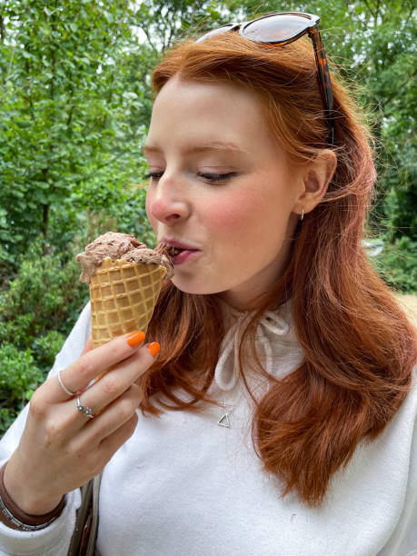 Close-up image of attractive redheaded woman wearing sunglasses on head, orange nail polish, holding waffle cone containing scoop of chocolate gelato with chocolate flake, eating crumbling chocolate, melting ice cream, woodland background, profile view Stock photo showing close-up, profile view of unrecognisable person holding a waffle cone with a scoop of chocolate ice cream and chocolate flake whilst walking in woodland. licking ice cream cone stock pictures, royalty-free photos & images