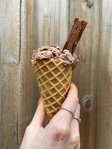 Stock photo showing close-up view of unrecognisable person holding a waffle cone with a scoop of chocolate ice cream and chocolate flake against a wooden fence background.