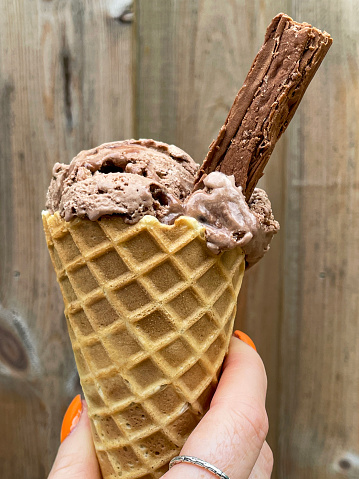 Stock photo showing close-up view of unrecognisable person holding a waffle cone with a scoop of chocolate ice cream and chocolate flake against a wooden fence background.