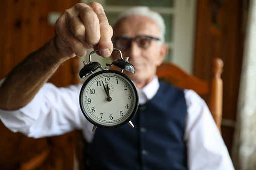 Senior man sitting in his armchair as he holds a small alarm clock.