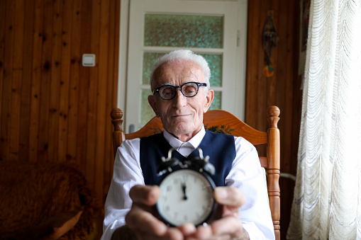 Senior man sitting in his armchair as he holds a small alarm clock.