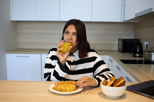 A young girl is having breakfast in the kitchen: drinking freshly squeezed orange juice and eating a croissant.