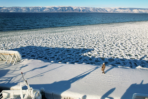 Frozen pier, ice floes and snow near the shore, water in Lake Baikal in December, aerial view. A woman admires a beautiful view. Beautiful winter landscape.