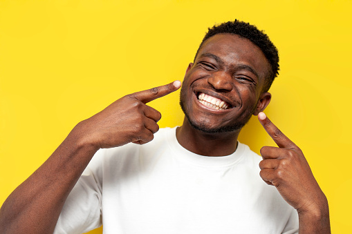 happy african american man pointing fingers at white toothy smile on yellow isolated background, dental care and whitening concept