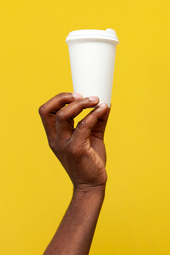 white paper cup of coffee in the hand of african american man, the guy holds empty packaging for drink on yellow isolated background, close-up